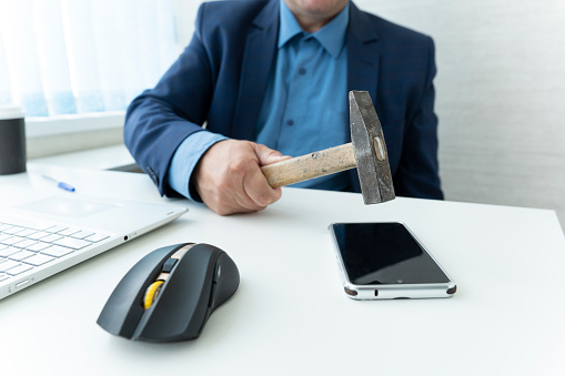 a businessman sitting in a table with a hammer in his hand ready to crash it on a smartphone