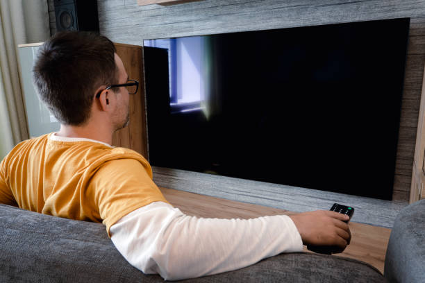 istock Man watching television, turning on plasma flatscreen TV set, pointing remote control at empty TV screen on white wall. Guy switching channels at home, back view. Mockup 1438023346