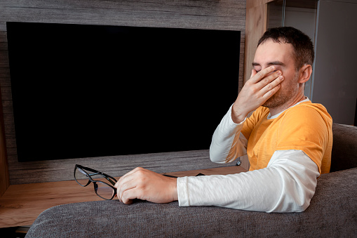 man feeling tired, touching eyes when watching tv at home. fatigue of vision . A viewer in a yellow shirt on the background of the TV