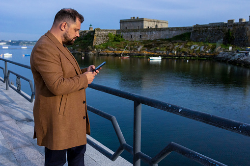 A man with a beard in a brown coat looks at his phone on the waterfront. A man in dark blue pants stands near the railing. In the background is an ancient castle in the ocean. Blue clear sky. There are small boats in the ocean. Attentiveness. Concentration. Thoughts. Business.