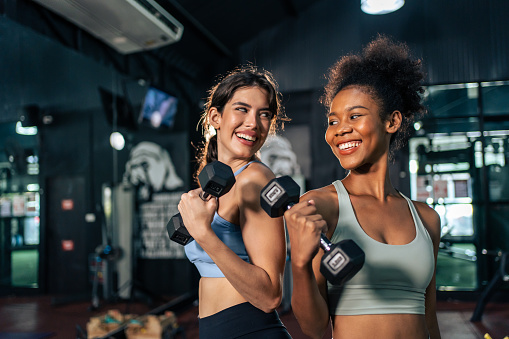 Latino and African sport woman exercising and build muscle in stadium. Active strong beautiful fit athlete girls in sportswear workout lifting weight dumbbell for arm and health care at fitness club.