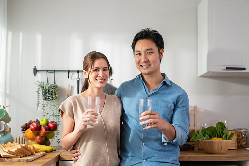 Asian attractive couple drinking a glass of water in kitchen at home. Young thirsty man and woman holding clean mineral natural in cup after waking up and sip in morning for health care in house.
