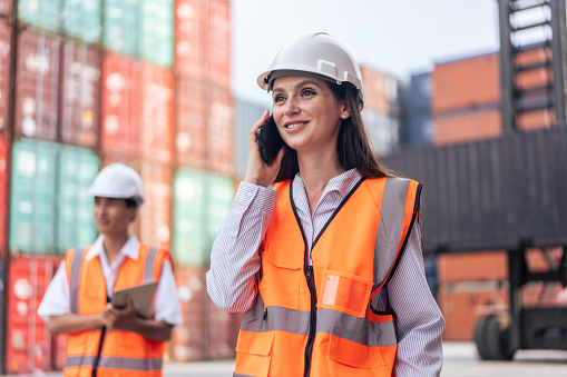 Caucasian businesswoman talk on phone while work in container terminal. Attractive young girl engineer process order and product at warehouse logistic in cargo freight ship for import export in harbor