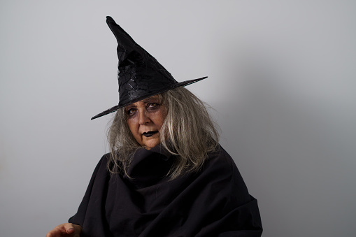 Cute senior woman, with long silver hair, dressed as a witch for Halloween, she is wearing a big witch hat. She is looking away from the camera. Defocused background with a wave pattern and some slight pink lighting.