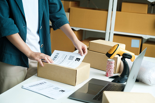 Young small business entrepreneur woman, online store onwer with shelves of cardboard boxes in the background packing product in mailing box, labeling delivery package for shipping to customer. Online shopping, e-commerce concept.