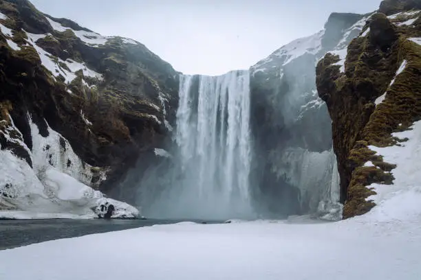 Skogafoss Waterfall in Winter under snowy overcast cloudscape. Falling water of the Skoga River. Snow and Ice covered Landscape of Skógafoss Waterfall. The famous Skógafoss is one of the biggest waterfalls in Iceland, with a width of 25m and a drop of 60m. Skógar, Rangárþing eystra, Southern Iceland, Sudurland, Iceland, Nordic Countries, Northern Europe