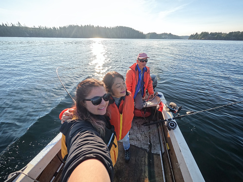 Asian mother embracing Eurasian daughter taking selfie of family trolling for salmon in Barkley Sound, British Columbia, Canada.