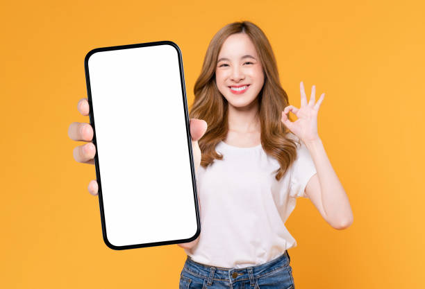Cheerful beautiful Asian woman holding smartphone and shows ok sign on light yellow background. stock photo