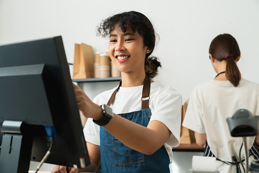 Happy young Asian woman cashier wears an apron and using pos terminal to input orders on coffee shop counter.