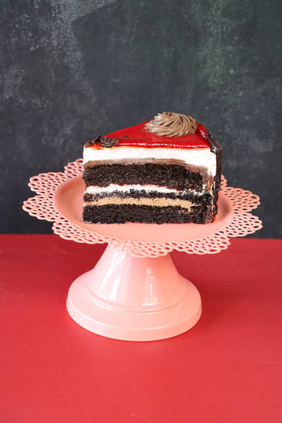 Stock photo showing close-up view of a slice of chocolate and cherry cake displayed against a mottled grey and red background. imagen de rebanada de chocolate y pastel de cereza, tres capas de bizcocho de chocolate separadas por crema de mantequilla en un soporte de pastel rosa, roseta de glaseado, glaseado de cereza, fondo gris moteado y rojo, enfoque en primer plano