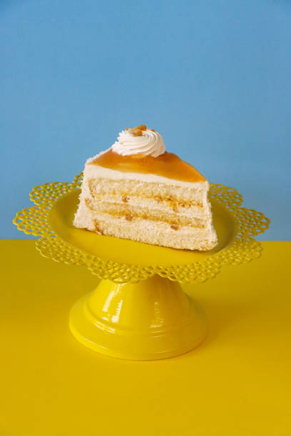 istock Close-up image of salted caramel cake slice, three vanilla sponge layers separated by vanilla buttercream and salted caramel on yellow cake stand, piped icing rosette, salted caramel glaze, yellow and blue background, focus on foreground 1438071974