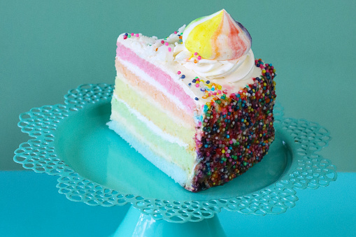 Stock photo showing close-up view of a slice of rainbow cake displayed against a turquoise blue background.