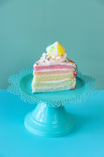 istock Image of rainbow cake slice, five rainbow sponge layers separated by vanilla buttercream on turquoise cake stand, piped icing rosette, rainbow meringue kiss, multicoloured hundreds and thousands, turquoise blue background, focus on foreground 1438071987