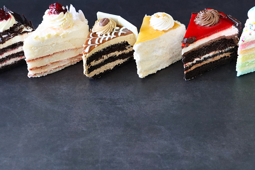 Stock photo showing close-up view of slices of Black Forest gateau, angel food cake, coffee and marbled gateau, salted caramel cake, chocolate and cherry cake and rainbow cake displayed in a row on a mottled black background.