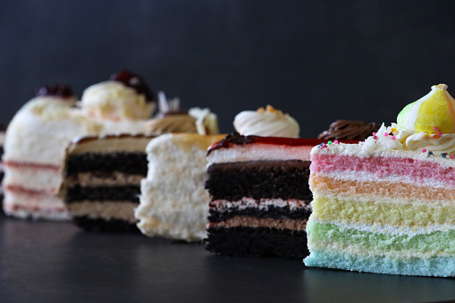 Stock photo showing close-up view of slices of Black Forest gateau, angel food cake, coffee and marbled gateau, salted caramel cake, chocolate and cherry cake and rainbow cake displayed in a row on a mottled black background.