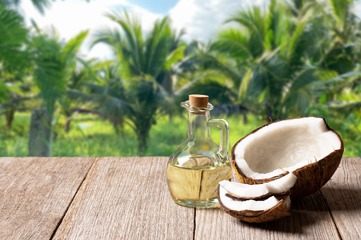 Coconut oil in glass bottle and brown coconut fruit on wooden table with coconut tree background.