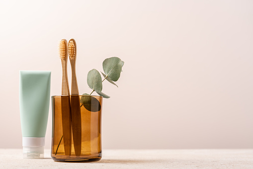 Bamboo toothbrushes in glass and tube of toothpaste on light background