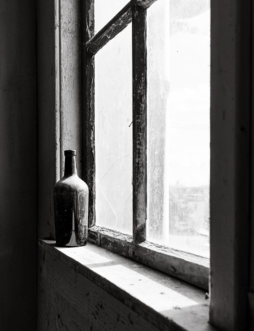 A vertical grayscale shot of an aged glass bottle covered in dust placed on the windowsill