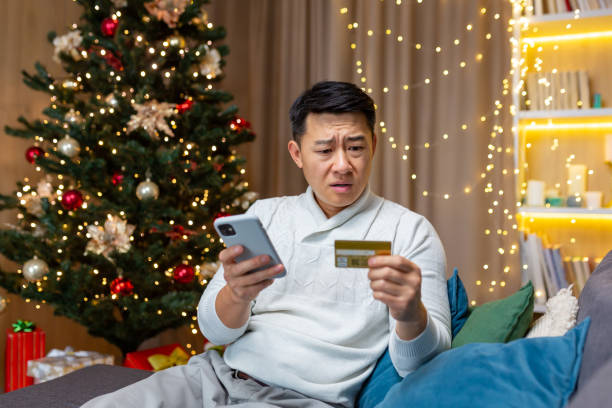 Dissatisfied Asian man trying to make a purchase in an online internet store, man cheated on Christmas, sitting on sofa in living room with smartphone in hands and credit card at home near Christmas tree. hombre asiático insatisfecho tratando de hacer una compra en una tienda en línea de internet, hombre engañado en navidad, sentado en el sofá de la sala de estar con teléfono inteligente en las manos y tarjeta de crédito en casa cerca del árbol de na