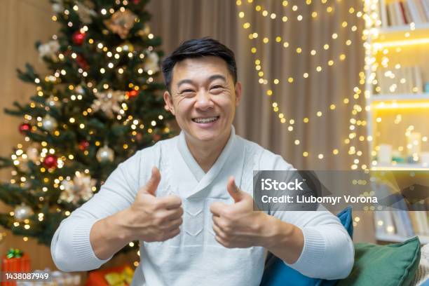 クリスマスのアジア人男性はウェブカメラを見て微笑み、指を肯定的に見せ、男は新年のクリスマスツリーの近くのソファに家で座っている