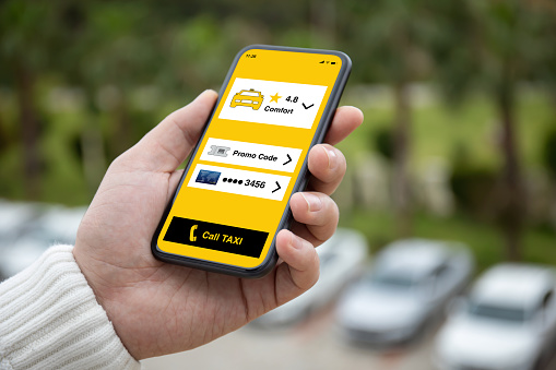 man hand holding phone with app call taxi on screen background white cars in car park