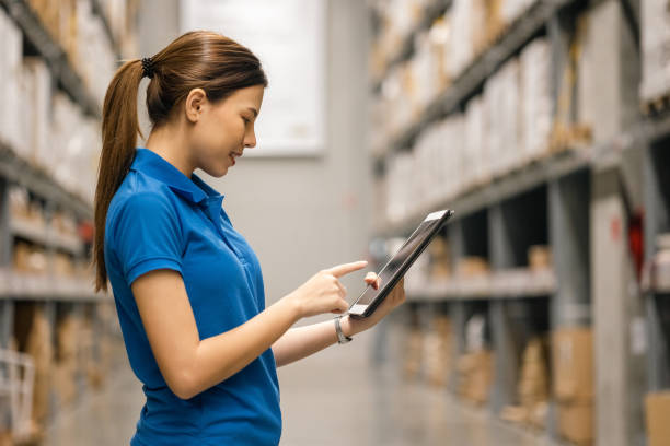Young female worker in blue uniform checklist manage parcel box product in warehouse. Asian woman employee holding tablet working at store industry. Logistic import export concept. joven trabajadora con uniforme azul de lista de verificación para administrar el producto de la caja de paquetes en el almacén. empleada asiática sosteniendo tabletas trabajando en la industria de tiendas. concepto logístico de importación y exportaci