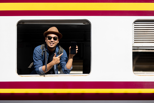 Young asian man travel by train. Sticking his head outta the train window showing smartphone. Backpacker arrival and departure at platform railway. Freedom  trip on vacation time holiday weekend.