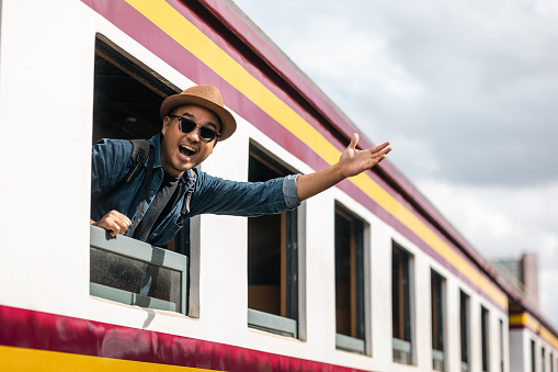 Young asian man travel by train. Sticking his head outta the train window. Explorer Backpacker arrival and departure at platform railway. Freedom  trip on vacation time holiday weekend.
