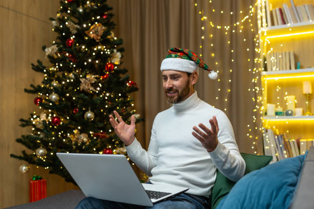 istock Man celebrating christmas and new year at home alone, using laptop for video call and remote communication with friends online, sitting on sofa in living room near tree, happy in hat 1438091009