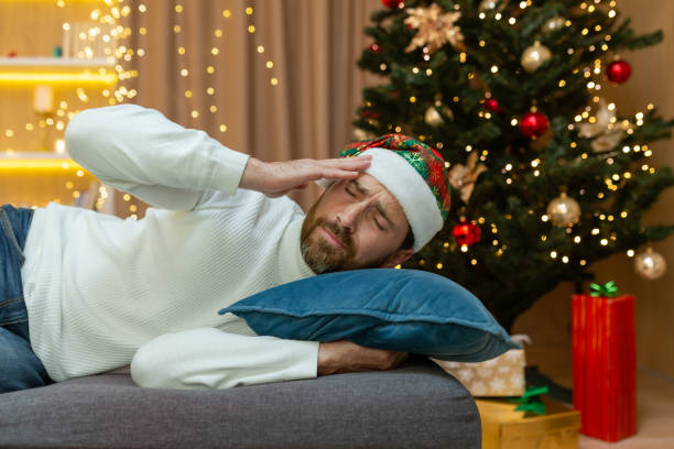 a man is tired for christmas, in a new year's hat, he is sleeping by the christmas tree on the sofa at home in the living room after celebrating christmas, he has a bad headache from a hangover - ressaca imagens e fotografias de stock