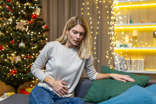 mujer con dolor abdominal y náuseas en la víspera de año nuevo sentada en casa en el sofá contra el fondo de un árbol de navidad. intoxicación, acidez estomacal, síndrome de dolor menstrual se siente mal - comer demasiado fotografías e imágenes de stock