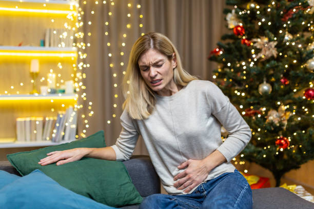 una mujer enferma se sienta sola en casa, tiene un fuerte dolor de estómago en navidad cerca del árbol de navidad en las vacaciones de año nuevo - christmas eating nausea illness fotografías e imágenes de stock