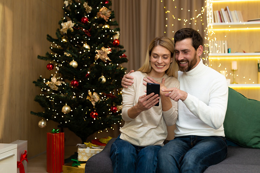 Married couple choose Xmas gifts on holiday online offers. Happy loving spouses sit by glowing New Year tree .