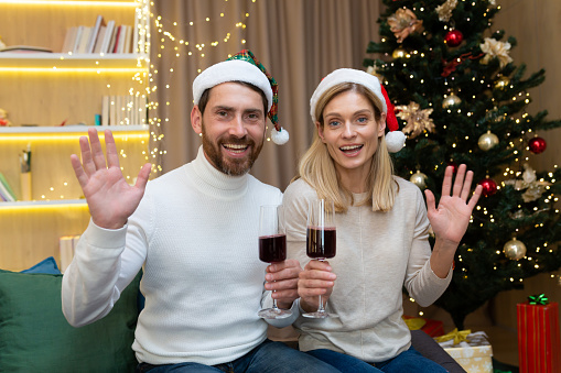 Mature married couple man and woman celebrating Christmas and New Year together, looking at web camera and smiling talking to friends on video call holding wine glasses in hands waving greeting home
