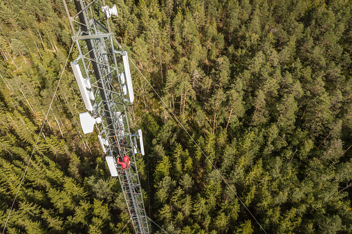 Working at height. Technician climbs up on a communications tower and installing the new 5G, LTE antenna in the middle of forest. Drone point of view.
