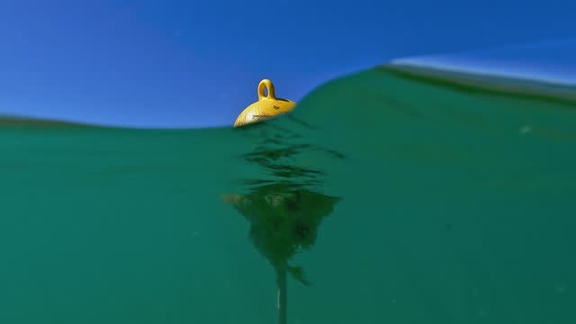 Half underwater tilt up view from seafloor to surface of yellow floating mooring buoy on sea water. Slow motion