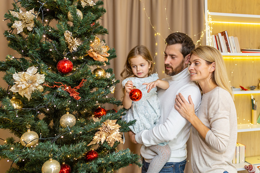 A young happy family decorates a Christmas tree for the Christmas holidays. Dad holds his daughter in his arms, the girl hangs a toy, mom hugs dad.
