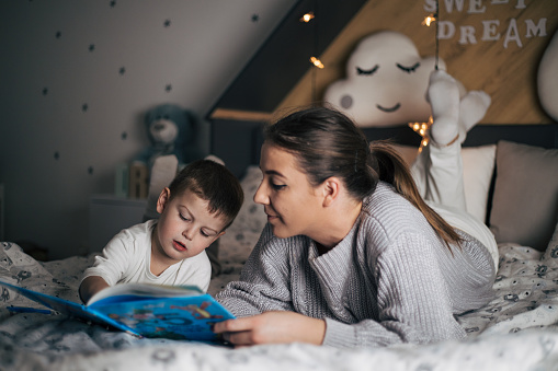 Mother and son in beautiful and loving relationship. Enyojing on bed playing and reeding books. Happiness and delight environment. Baby boy bed with toys, big pillows and lights.