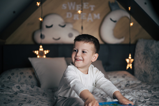 Young boy sitting on bed with toys and star shaped lights.  Love and sweetness can be seen in his eyes. He is happy and ready to hear a story before sleep in his bedtime. Big cloud and moon shapped pillows behind him on the wall with ?*sweet dream* sign letters.