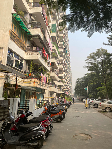 Noida, Uttar Pradesh, India  - November 1, 2022: Stock photo showing street in Noida, India in urban area passing apartment block with rows of parked cars and motorbikes. Apartment complexes are popular in Indian as flats are more economical to build for property developers.