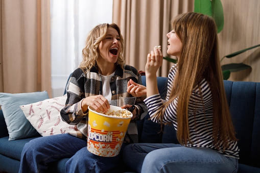 Two happy women have fun sitting on the couch in the apartment and feeding each other popcorn