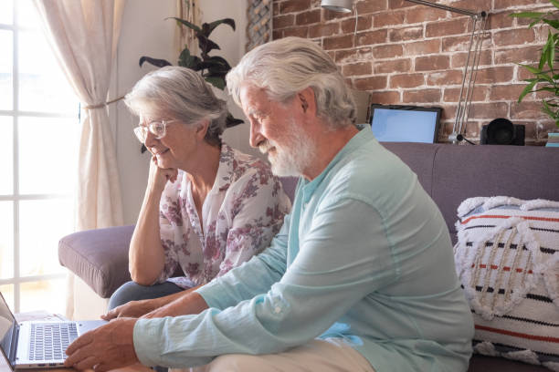 istock Two caucasian mature modern senior people sitting on sofa at home using laptop computer. Elderly couple gray haired relaxing and smiling together looking at same laptop. Brick wall background 1438123682