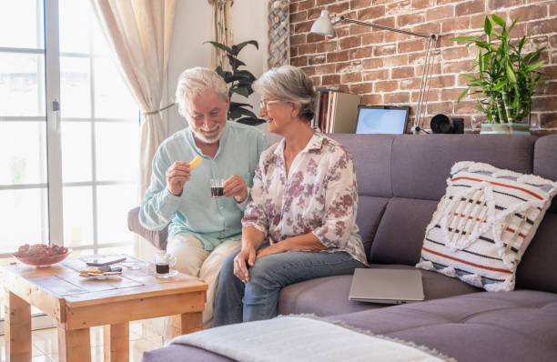 istock Two caucasian senior people sitting on sofa at home drinking a coffee. Elderly couple gray haired relaxing and smiling having a break. Technological devices around them. Brick wall background 1438123729