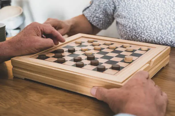 Photo of Two senior people at home while playing a game of checkers on wooden table.