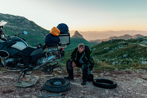 A concerned motorbike rider is sitting on the road in the middle of nowhere with his head in hand. He tried to change the tire but he failed