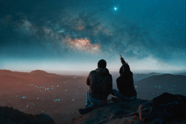silhouette of a couple sitting on top of a hill looking at the stars over the city stock photo