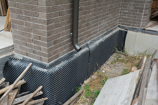 House foundation basement waterproofing, damp proofing with rigid insulation. Installation of a dimpled waterproofing membrane on the exterior basement wall and rain gutter downspout drain pipe.