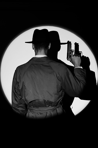 A dark silhouette of a male detective in a coat and hat with a gun in his hands in the noir style. A dramatic portrait in the style of detective films of the 1950s. The silhouette of a spy in a circle of light. monochrome