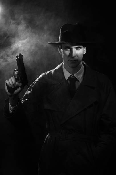 une silhouette sombre d’un homme en imperméable et chapeau avec un pistolet à la main dans le style du crime noir. un portrait noir dramatique dans le style des détectives des années 1950. monochrome - police crime gun gang member photos et images de collection