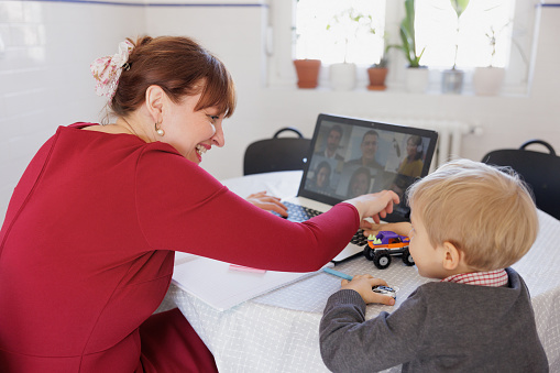 Woman sitting at the table, using laptop for video conference, smiling and scolding her toddler for distracting her